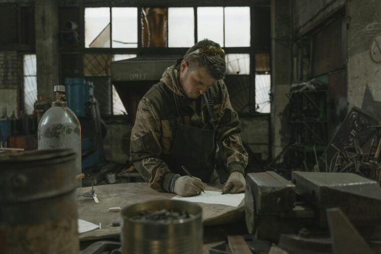 A metalworker in camouflage jacket sketches designs in an industrial workshop.
